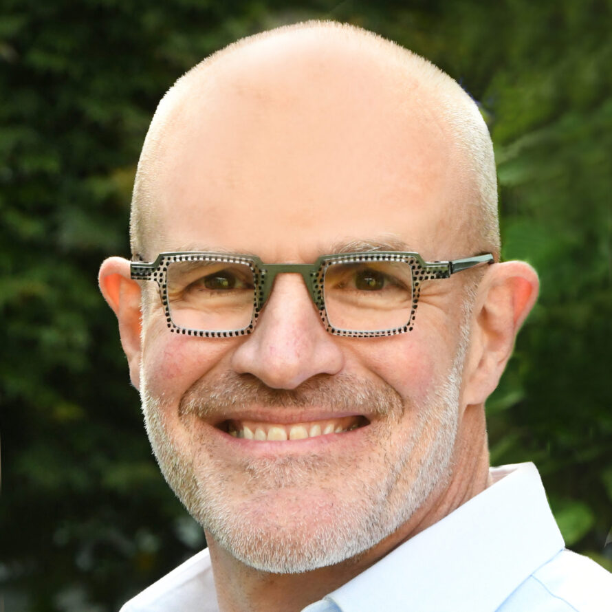 Portrait of Jay Keasling, a person with short gray beard, wearing green rectangular glasses and light shirt with top bottoms open.