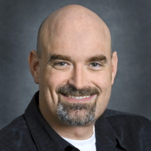 Portrait of Blake Simmons, a bold person with black and gray goatee, wearing a dark shirt over a white t-shirt. Photographed in front of a dark backdrop.