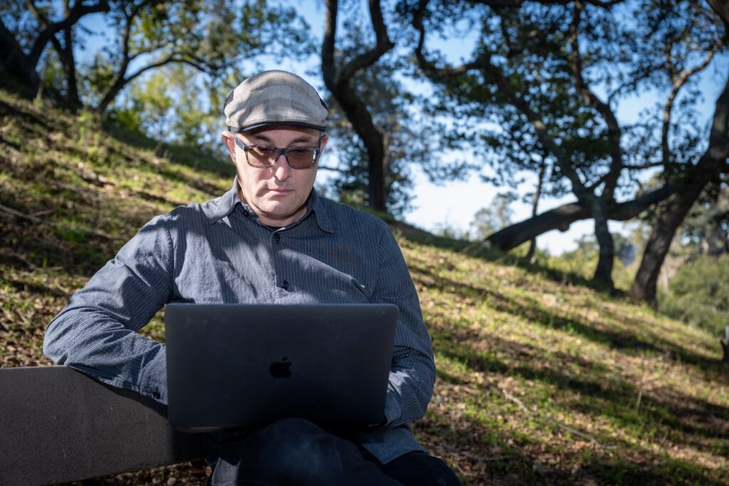 Dylan Chivian works at a computer while sitting outside on a sun-dappled hillside with with oak trees.