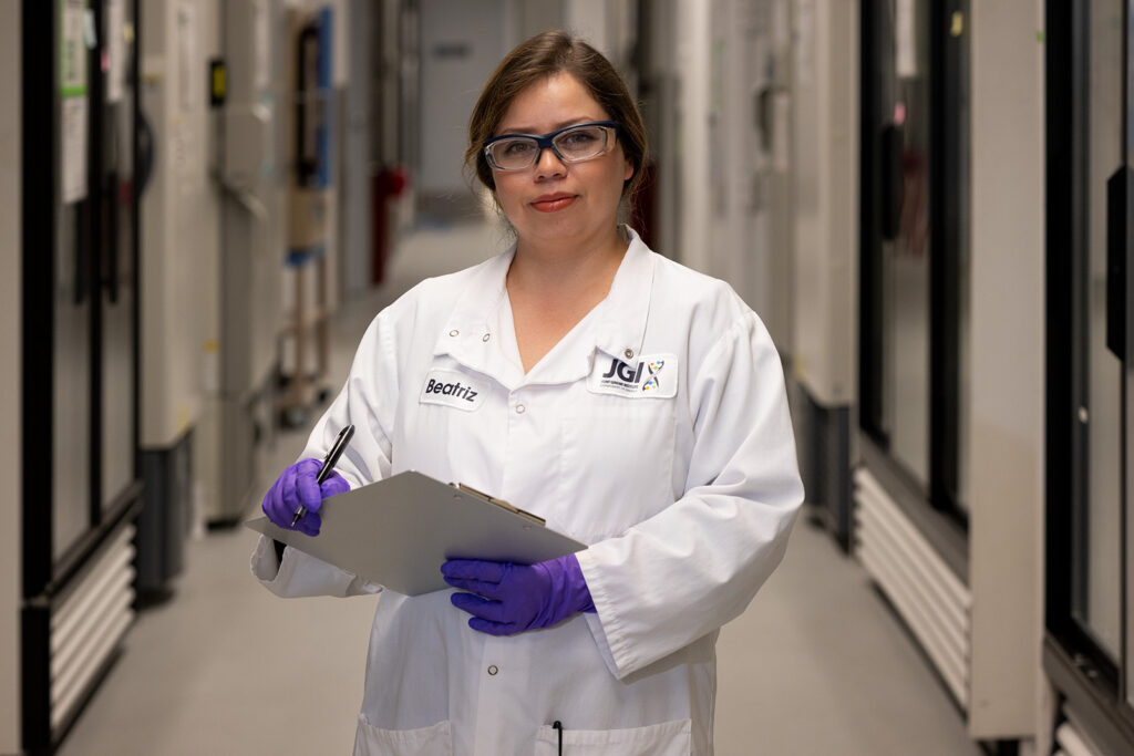 Portrait of Beatriz Rett wearing a lab coat, safety goggles, and latex gloves and carrying a clipboard inside the Integrative Genomics Building at Berkeley Lab.