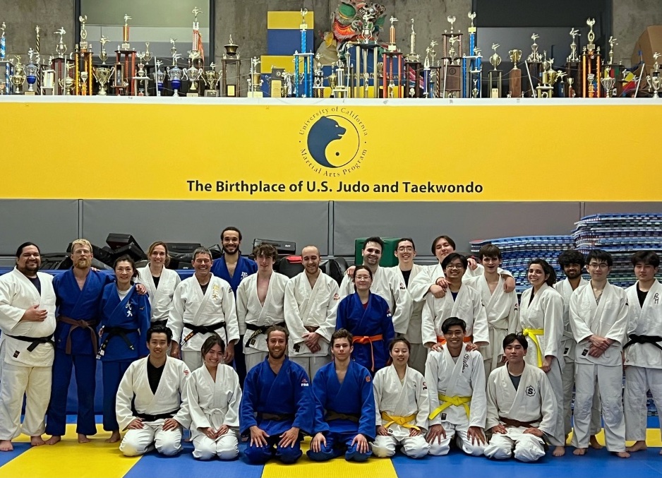 Group photo of Lorenzo Washington's Dojo Community, posing in a gym with an array of trophies in the background. A banner above the group reads: "The Birthplace of U.S Judo and Taekwondo."