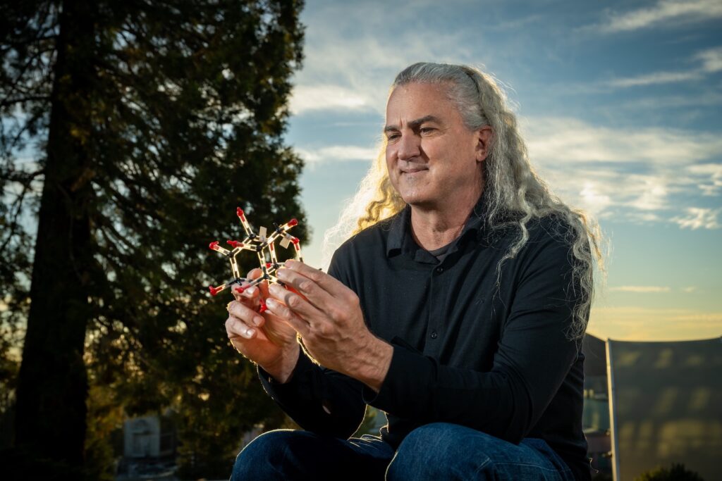 Photo of Nigel Moriarty smiling while holding a model of a molecule against the backdrop of a vivid sky at sunset.