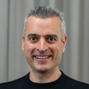 Portrait of Nathan Hillson, a person with short gray and dark hair, smiling and wearing a black t-shirt. Photographed in front of a gray curtain backdrop.