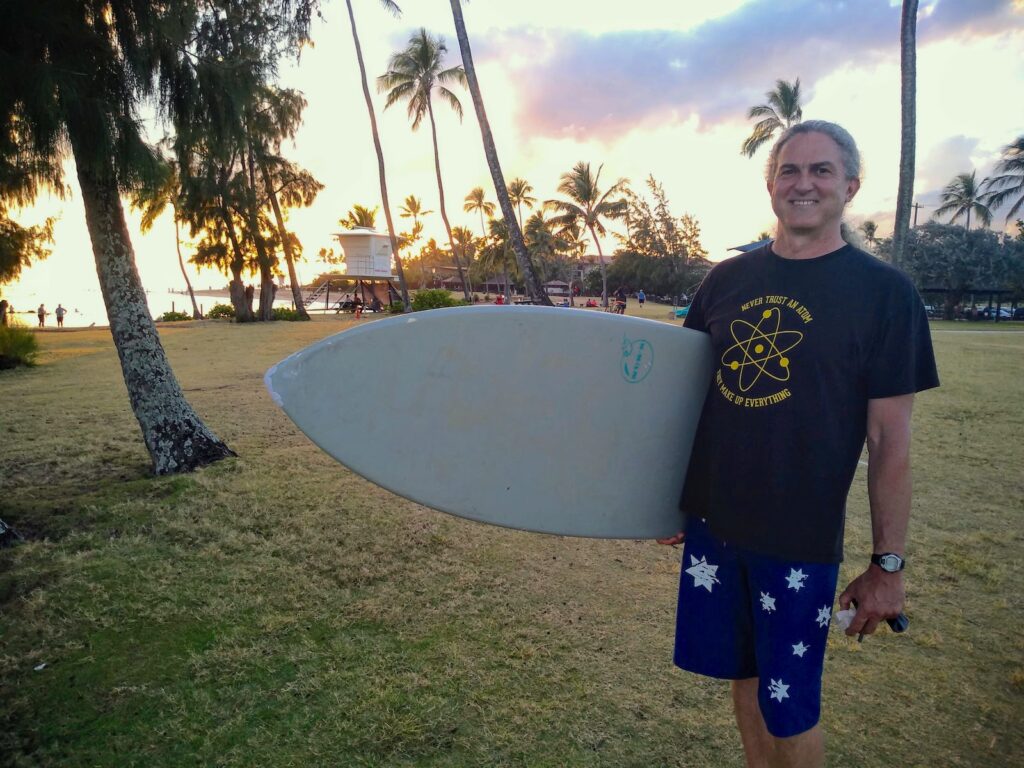 Nigel Moriarty poses for a photo while carrying a sufboard. In the background, a sunset sky above a tropical beach is visible.