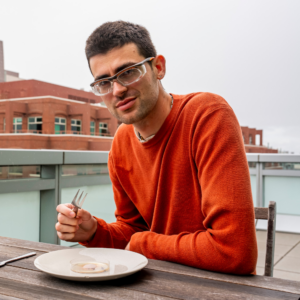 A man wearing an orange sweater and laboratory goggles is pictured on an outdoor balcony. He is holding a fork and has a plate in front of him that contains a petri dish of mold.