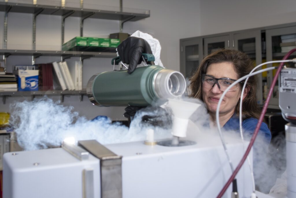Female scientist smiles while pouring liquid nitrogen from a canister  to the Fourier-Transform Infrared Spectroscopy (FTIR) device. 