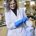 Scientist wearing a lab coat, safety goggles, and gloves holds open a canister of liquid nitrogen.