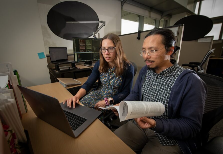 A woman with shoulder-length brown hair and glasses works at a computer while a man of Asian descent with black hair, beard, and mustache and wearing glasses references a journal article.