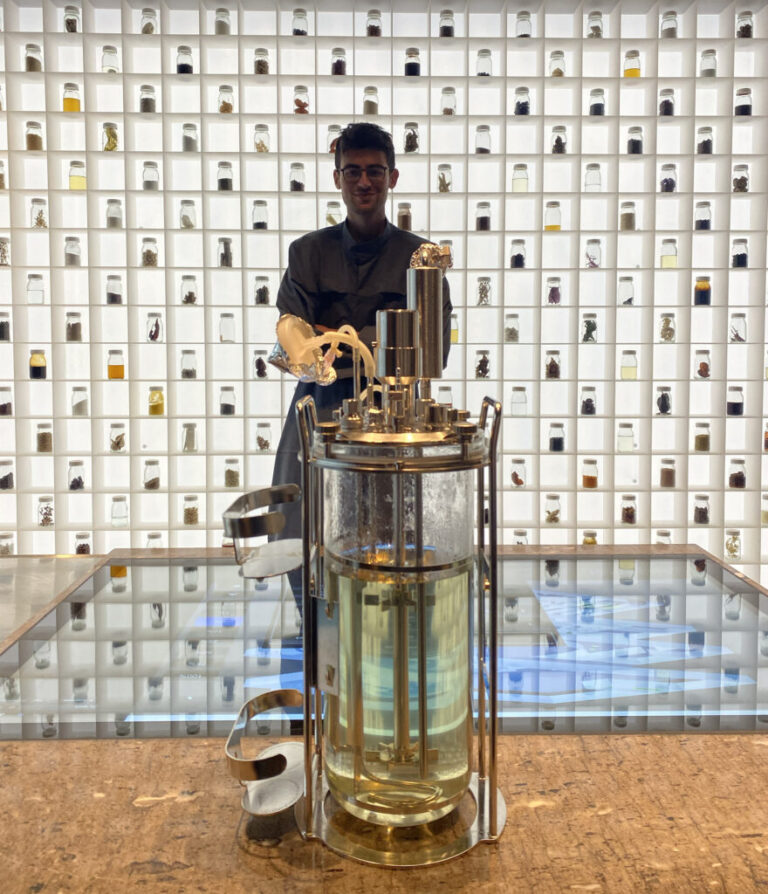 A dark-haired man in glasses stands next to a waist-high glass container of fluid and in front of a lighted wall of jars