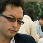 In a dark blazer and white button down shirt with slightly tinted glasses, Stephen Chan looks intently at the camera with plans in the distance behind him. In the background on the right side of the photo are several people.