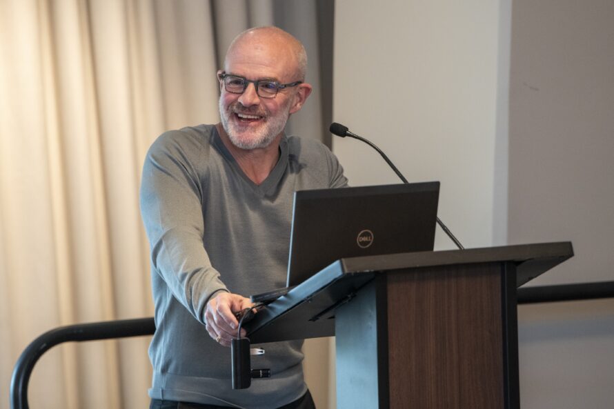 Jay Keasling, a bald man with a grey beard, wearing black framed glasses and a grey sweater, stands at a wooden podium during a presentation.