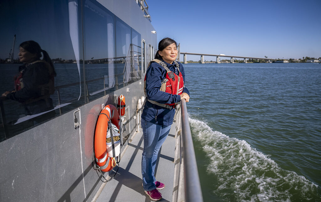 Research scientist Lauren Lui smiles while looking out at the blue-green waters of the Sacramento-San Joaquin River Delta from the deck of a boat. The Antioch bridge is visible in the distance.
