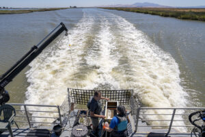 View from the deck of the research vessel Sentinel looking out over one of the San Francisco Estuary’s wide open channels near the Sacramento-San Joaquin river Delta. Two field ecologists set up equipment in the foreground on the boat's deck.