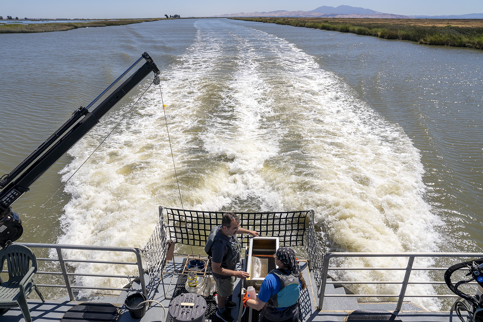 View from the deck of the research vessel Sentinel looking out over one of the San Francisco Estuary’s wide open channels near the Sacramento-San Joaquin river Delta. Two field ecologists set up equipment in the foreground on the boat's deck.