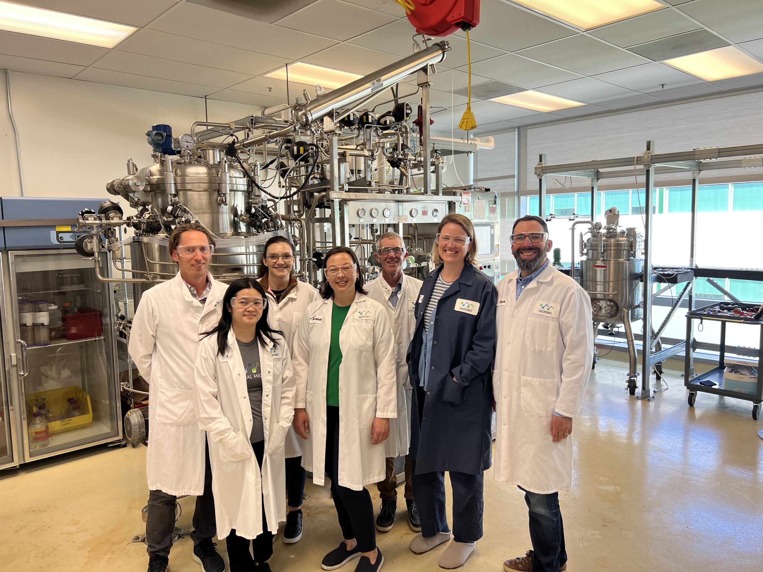 A group of researchers in lab coats stand in front of a fermenter.