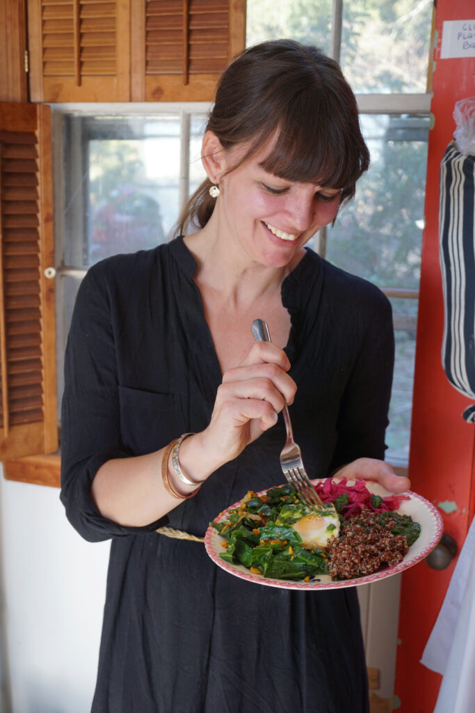 A woman smiles while taking a forkful from a prepared plate of colorful, fresh food.