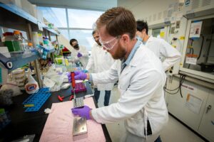 People in a lab wearing white lab coats perform an experiment with a pipette and tray.