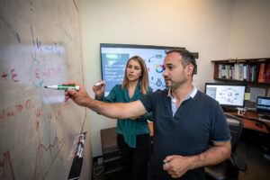 A woman and a man discuss equations written on a white board.