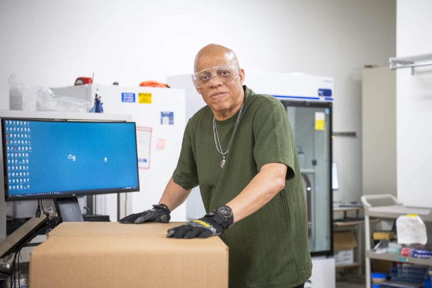 A person in a green shirt and protective gloves stands beside a cardboard box in a lab, with a monitor and equipment in the background.