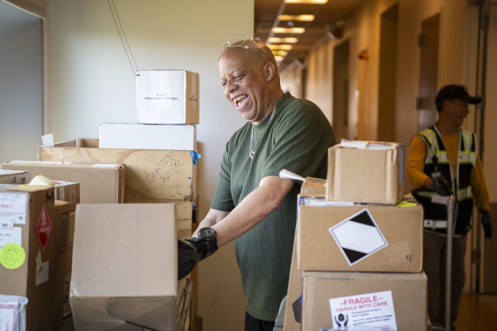 A man smiling while sorting cardboard boxes in a hallway, with another person in a safety vest in the background.