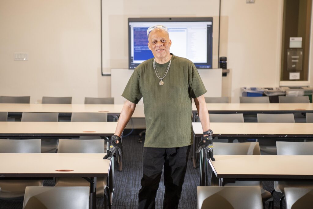 Person standing in a conference room, touching desks, with a screen and desks in the background.