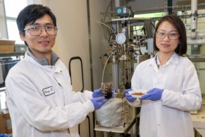 Two researchers in a laboratory setting wearing white lab coats and standing in front of various equipment hold samples of brown-colored lignin.