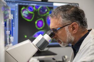 Man looking into a microscope with multi-colored cells on a display behind him.