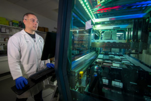 an individual in safety googles, lab coat and rubber gloves stands in front of scientific equipment