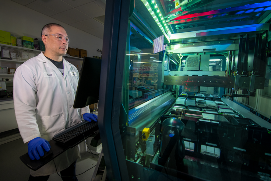 an individual in safety googles, lab coat and rubber gloves stands in front of scientific equipment