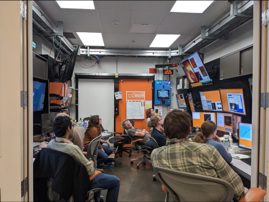 People seated in a laboratory control room surrounded by computer monitors, control panels, and scientific equipment.